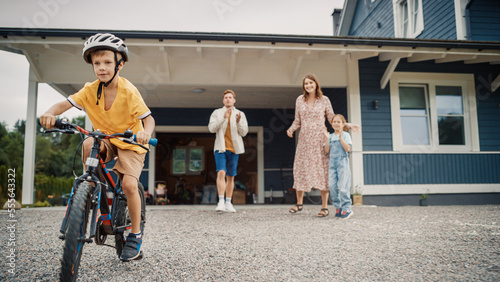 Cheerful Father Teaching His Son to Ride a Bicycle in The Front Yard of Their Beautiful Residential Area Home. Boy Wearing a Helmet, Balancing Himself on Bike. Family Cheering Him Up for His Success.
