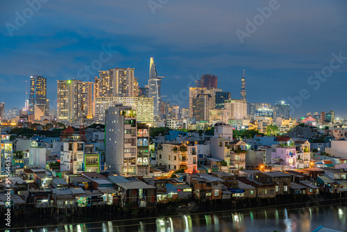 HO CHI MINH  VIETNAM - December 3  2022  Slum wooden house on the Saigon river bank  in front of modern buildings at night in ho chi minh city. View to district 1  see Bitexco tower  Landmark 81.