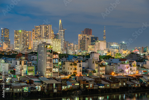 HO CHI MINH, VIETNAM - December 3, 2022: Slum wooden house on the Saigon river bank, in front of modern buildings at night in ho chi minh city. View to district 1, see Bitexco tower, Landmark 81.