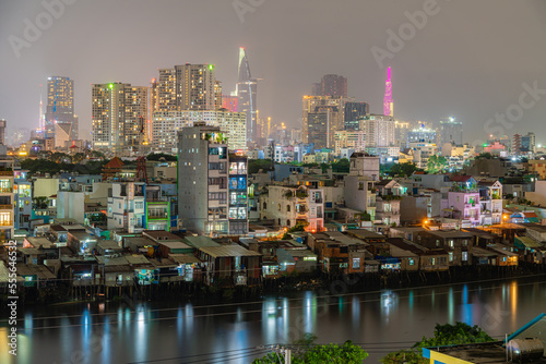 HO CHI MINH, VIETNAM - December 3, 2022: Slum wooden house on the Saigon river bank, in front of modern buildings at night in ho chi minh city. View to district 1, see Bitexco tower, Landmark 81.