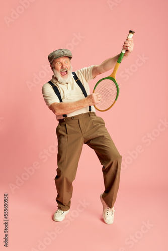 Portrait of senior, old man in stylish classical clothes posing with racket, over pink background. Illusion of guitar © master1305