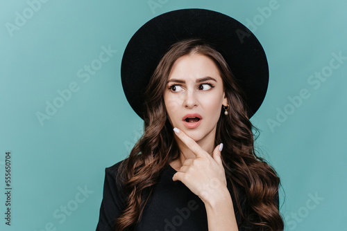 Great idea! Attractive hispanic young woman in black hat and black t-shirt with wide open eyes and mouth touches  her chin with surprised face. Solution was so simple. Look there, that's amazing.