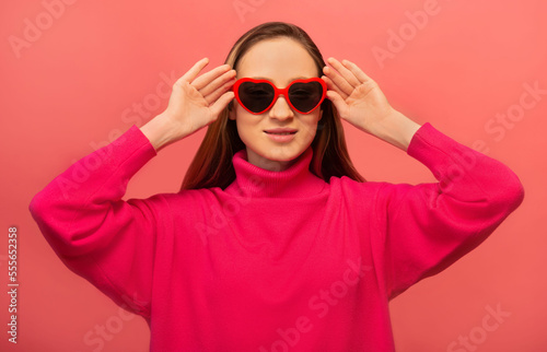 Portrait of happy smiling cheerful girl holding black and red heart shaped glasses and wearing pink sweater on pink background.St Valentines Day, International Womens Day or lifestyle concept.