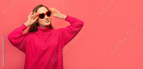 Stylish happy cheerful girl in warm pink dress with heart shaped glasses looking up on pink background with copy space.

St Valentines Day, International Womens Day or lifestyle concept. Banner.
