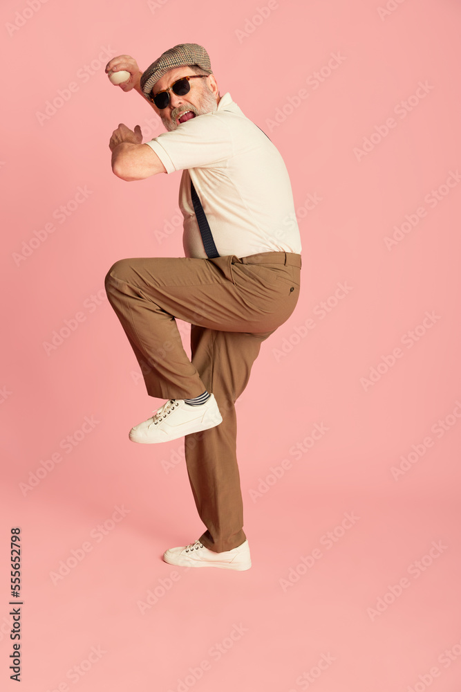 Fototapeta premium Portrait of senior man in stylish classical clothes posing, serving baseball ball over pink background. Sport club