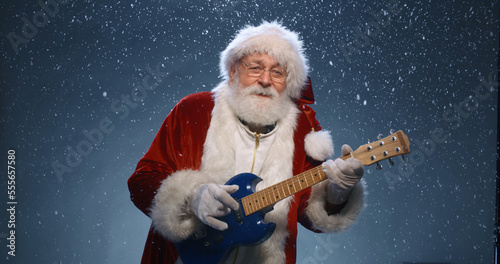 Portrait of cool Santa Claus in glasses playing the guitar and singing. Merry Christmas. Party. close up shot. Blue background with snowfall.