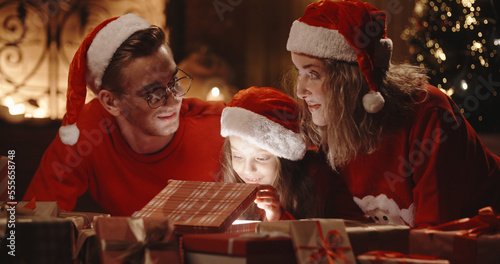 Caucasian family of three sitting in decorated room near christmas tree, little girl opening her christmas gift with something special - christmas spirit, togetherness concept 