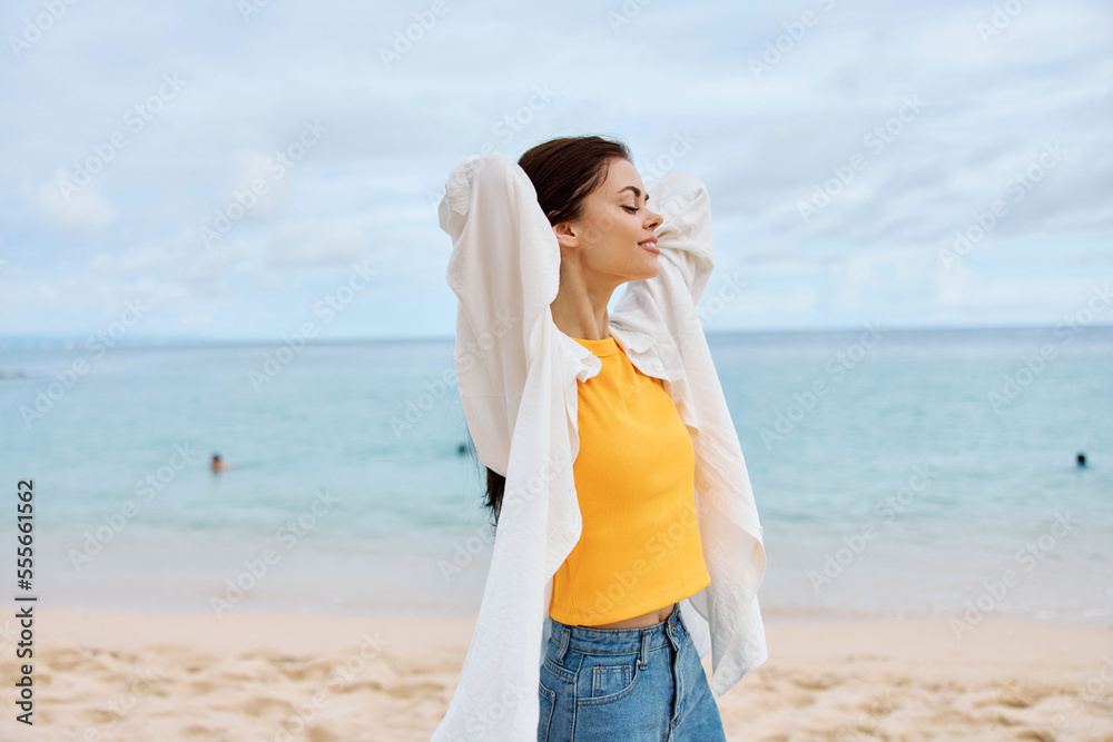 Happy woman smile with teeth with long hair brunette walks along the beach in a yellow t-shirt denim shorts and a white shirt near the sea summer journey and feeling of freedom, balance Happy woman smile with teeth with long hair brunette walks along the beach in a yellow t-shirt denim shorts and a white shirt near the sea summer journey and feeling of freedom, balance