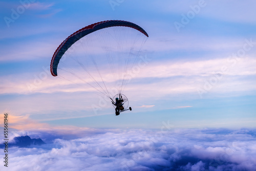 Silhouette of the paraglider with paramotor transportation control flying through soft sunlight fog white cloud blue sky.