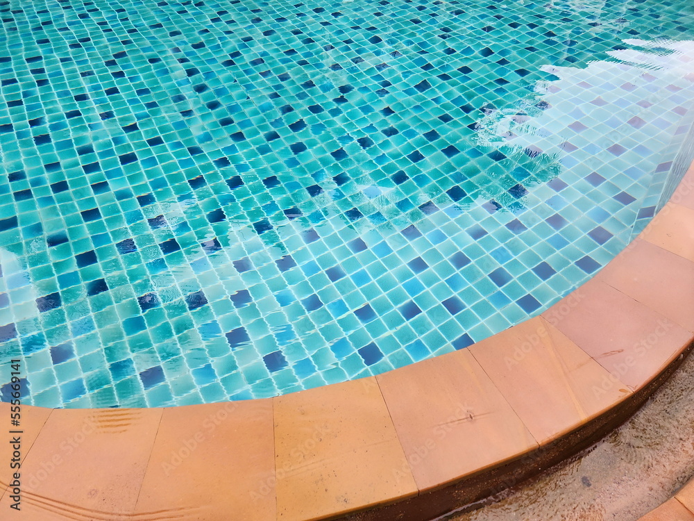 Water ripples in swimming pool with blue tile floor and brown pool edge. Turquoise blue pool, blue sky, ceramic mosaic tiles, various shades in the pool.
