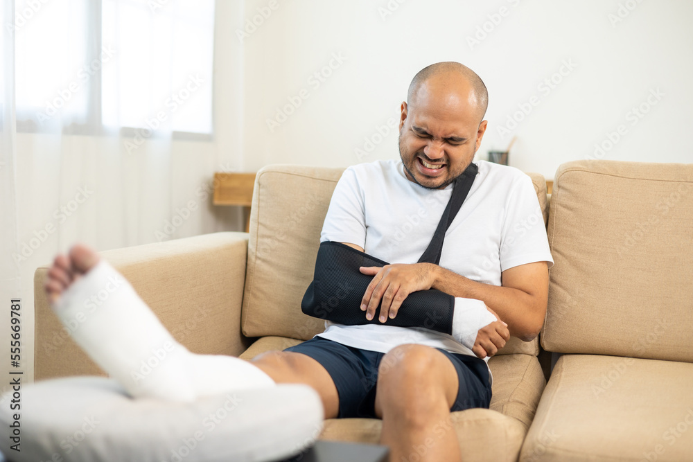 Man suffered pain from accident fracture broken bone injury with leg splints in cast neck splints collar arm splints sling support arm in living room. Social security and health insurance concept. Man suffered pain from accident fracture broken bone injury with leg splints in cast neck splints collar arm splints sling support arm in living room. Social security and health insurance concept.