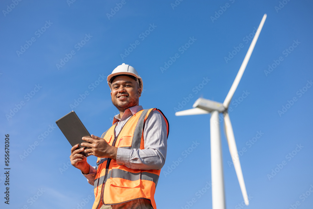 Engineer India man working with tablet at windmill farm Generating electricity clean energy. Wind turbine farm generator by alternative green energy. Asian engineer checking control electric power Engineer India man working with tablet at windmill farm Generating electricity clean energy. Wind turbine farm generator by alternative green energy. Asian engineer checking control electric power