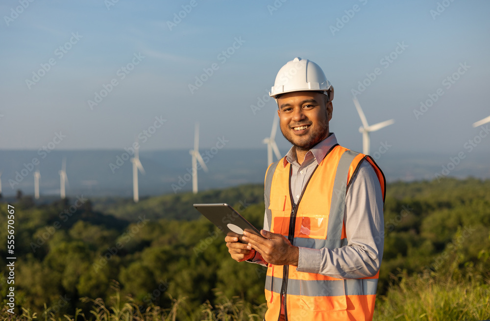 Fototapeta premium Engineer India man working with tablet at windmill farm Generating electricity clean energy. Wind turbine farm generator by alternative green energy. Asian engineer checking control electric power