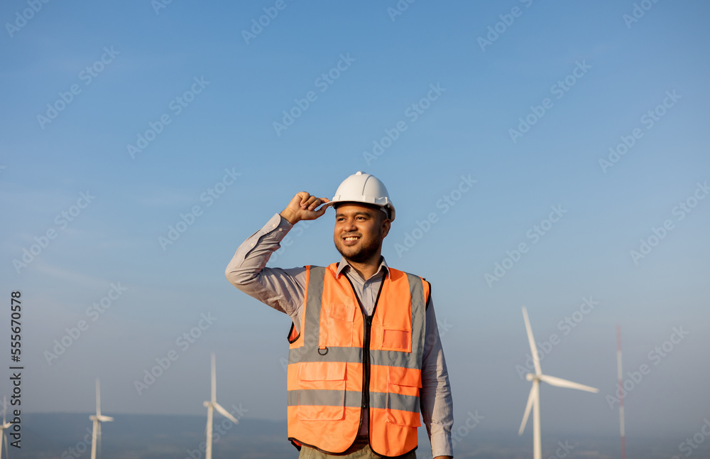 Engineer India man working at windmill farm Generating electricity clean energy. Wind turbine farm generator by alternative green energy. Asian engineer checking control electric power Engineer India man working at windmill farm Generating electricity clean energy. Wind turbine farm generator by alternative green energy. Asian engineer checking control electric power