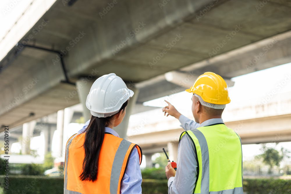 Two Asian worker engineer man and woman architect looking construction with white safety helmet in construction site. Standing at highway concrete road site. Work planning with blueprint and tablet. Two Asian worker engineer man and woman architect looking construction with white safety helmet in construction site. Standing at highway concrete road site. Work planning with blueprint and tablet.