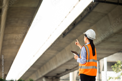 Asian engineer worker woman or architect looking construction with tablet wear white safety helmet in construction site. Standing at highway concrete road site. Progress planning of highway bridge.