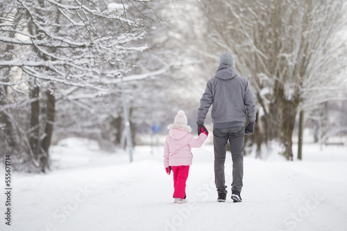 Little daughter and young adult father walking on white snow covered sidewalk at park. Spending time together in beautiful cold winter day. Enjoying stroll. Back view.