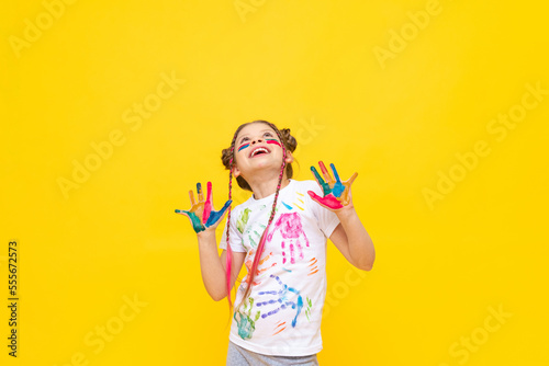 A little girl, painted with paints, looks at your advertisement on a yellow isolated background. The art of painting with paints for children. Art courses for schoolchildren. Copy space.