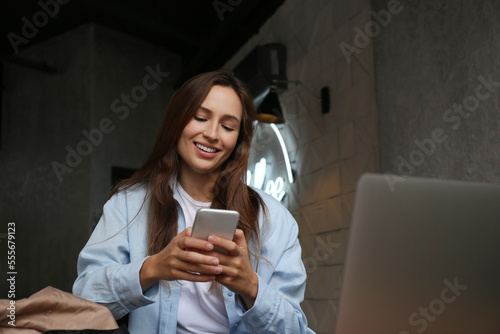 Portrait of young beautiful woman dressed in smart casual style sitting in a coffee shop with her laptop and checking her phone. Remote work concept. Close up, copy space, background.