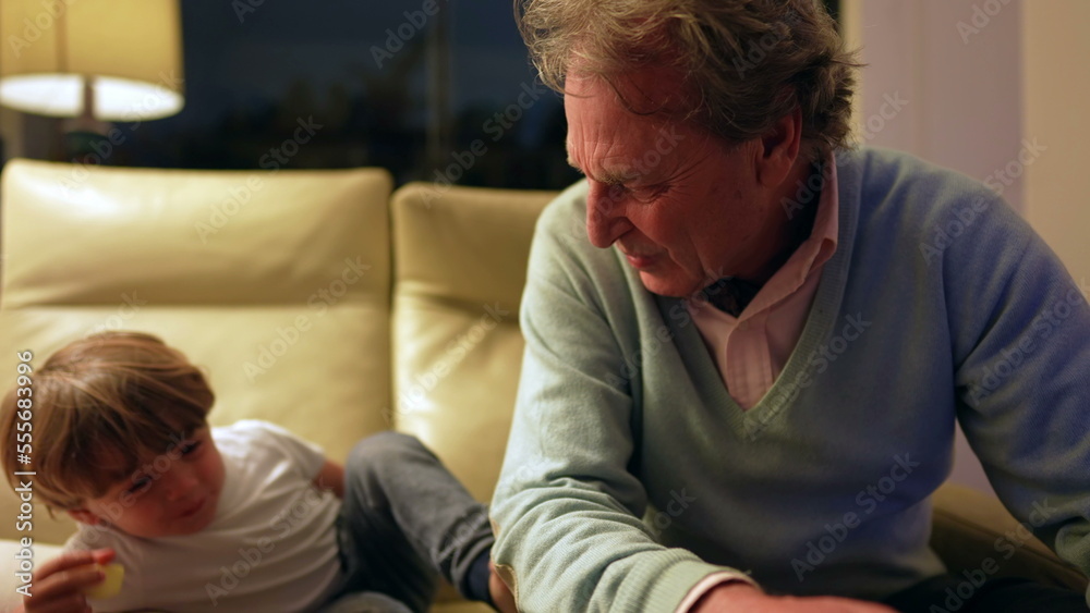 Grandfather and grandson hanging out together in living room couch. Authentic real life family generational relationship at night sitting on couch Grandfather and grandson hanging out together in living room couch. Authentic real life family generational relationship at night sitting on couch
