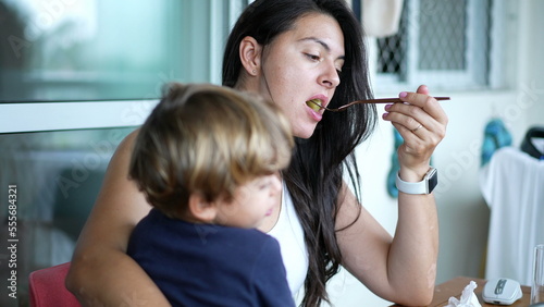 Mother eating while holding child in arms. Mom feeding small boy with fork