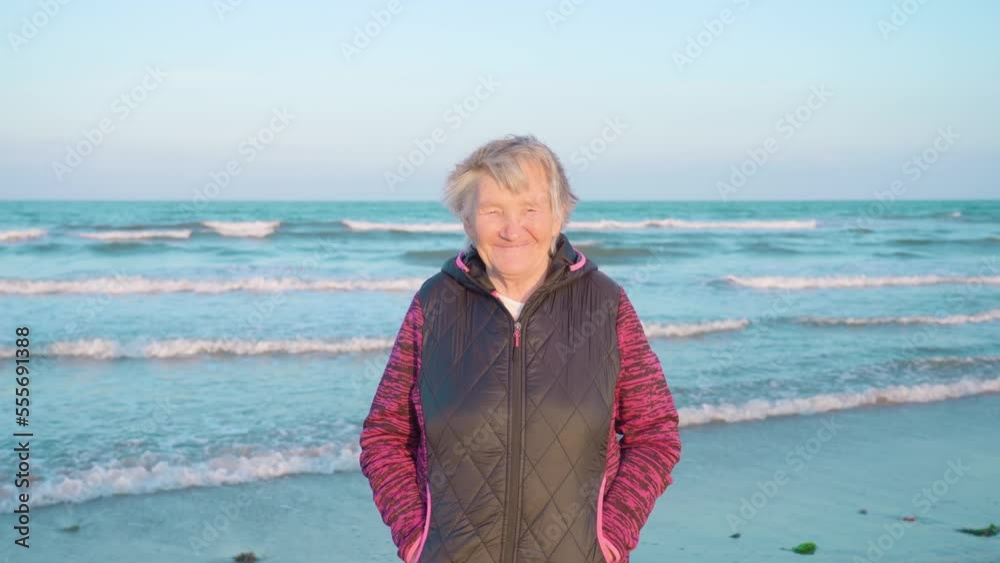 Old woman with short grey hair in warm windbreaker smiles happily standing with hands in pockets on azure sea beach closeup Old woman with short grey hair in warm windbreaker smiles happily standing with hands in pockets on azure sea beach closeup