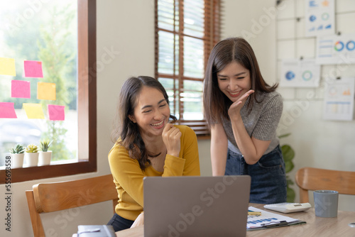 Two asian businesswoman expressing happiness in workplace smiling and raising hands in joy with laptop computer in modern office.