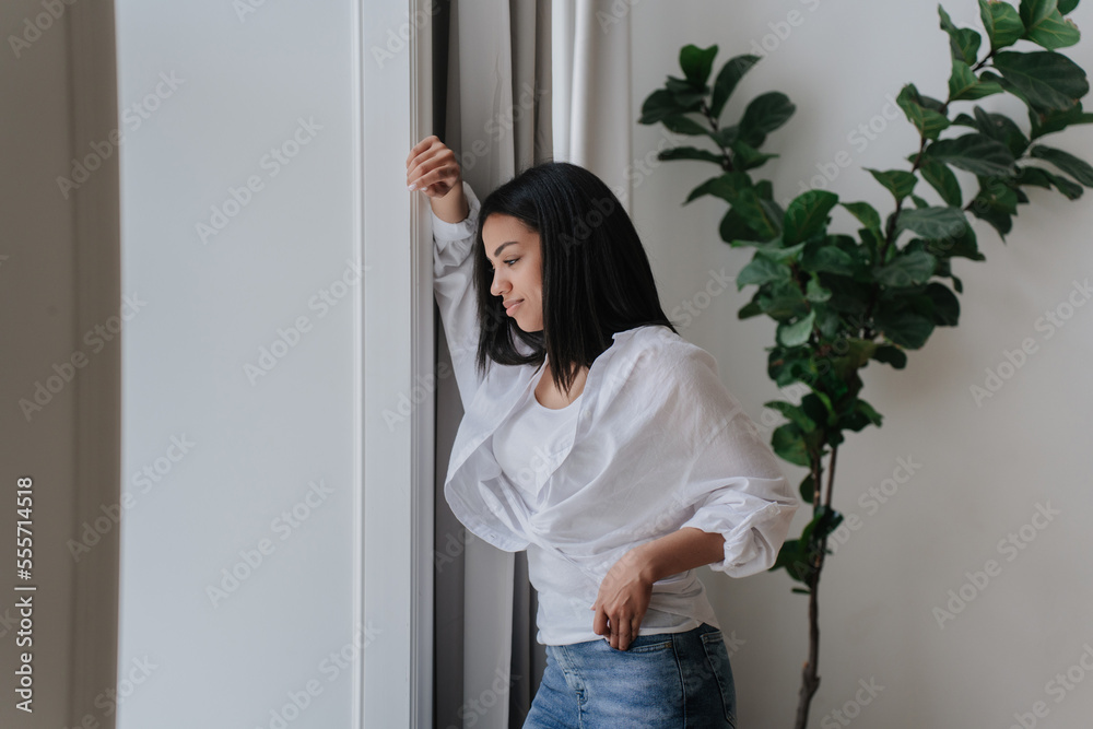 Satisfied African American young woman in white shirt and blue jeans standing at window with plant on background. Pretty Brazilian girl waits for boyfriend prepares for date. Beautiful people. Satisfied African American young woman in white shirt and blue jeans standing at window with plant on background. Pretty Brazilian girl waits for boyfriend prepares for date. Beautiful people.