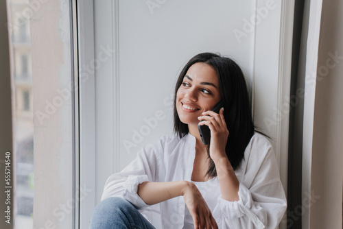 African American young woman in love talking by phone with boyfriend sitting on windowsill looking through window happy smiling. Brazilian girl in white shirt and blue jeans using cell phone at home.
