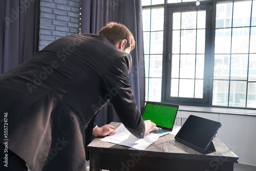 An unrecognizable businessman, he stands and types on his laptop using a chromakey on the background of the window. With a space to copy. High quality photo © daryakomarova