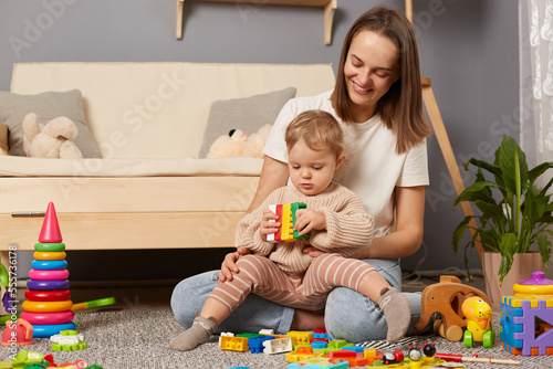 Horizontal shot of smiling dark haired woman in white t shirt sitting on floor with her baby daughter and playing with colorful blocks, spending free time for early development lesson.