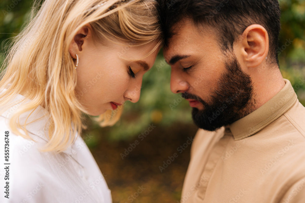 Close-up of loving married couple standing foreheads touched with closed eyes. Happy young bearded man and pretty blonde woman on romantic date in summer park. Couple relaxing heads embracing.