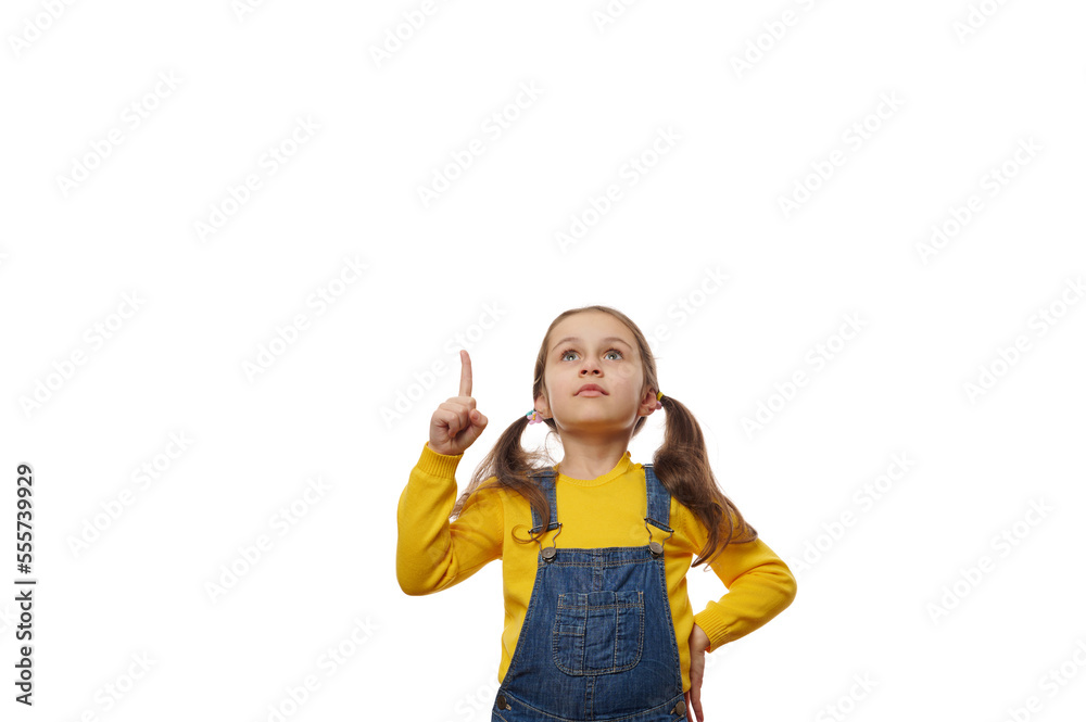 Caucasian lovely little girl with two ponytails, in bright yellow sweater and blue denim overalls, looks up and points forefinger at copy space on white background, for advertising promotional text Caucasian lovely little girl with two ponytails, in bright yellow sweater and blue denim overalls, looks up and points forefinger at copy space on white background, for advertising promotional text