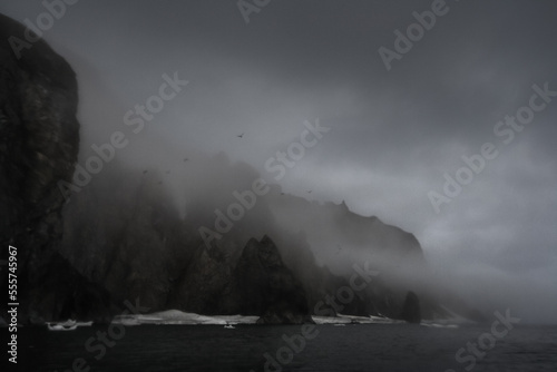 Summer night arctic landscape. July. Epic view of the rocky sea coast. Cloudy and foggy rainy weather. Ice floes near the shore. Coast of the Bering Strait, Chukotka Peninsula, Far East of Russia.