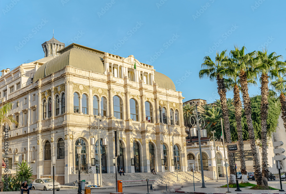 The national theater Mahiedine Bachtarzi side view in Square Port Said, Casbah. With palm trees in a sunny day with sunlight and shadows on the facade. Three unrecognizable people sitting on stairs.