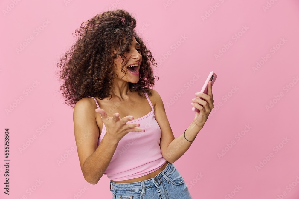 Woman blogger holding phone video call takes selfies, with curly hair in pink smile t-shirt and jeans poses on pink background, copy space, technology and social media, online Woman blogger holding phone video call takes selfies, with curly hair in pink smile t-shirt and jeans poses on pink background, copy space, technology and social media, online