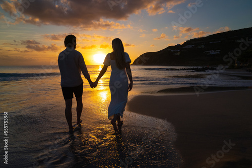 Young romantic dressed up couple walking down island beach towards the picturesque sunset