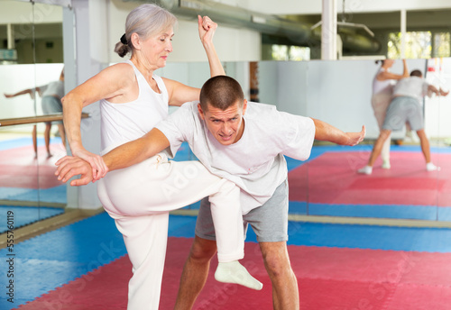 Concentrated elderly woman practicing self defense techniques, performing elbow and knee strikes with arm hold to male sparring partner in gym