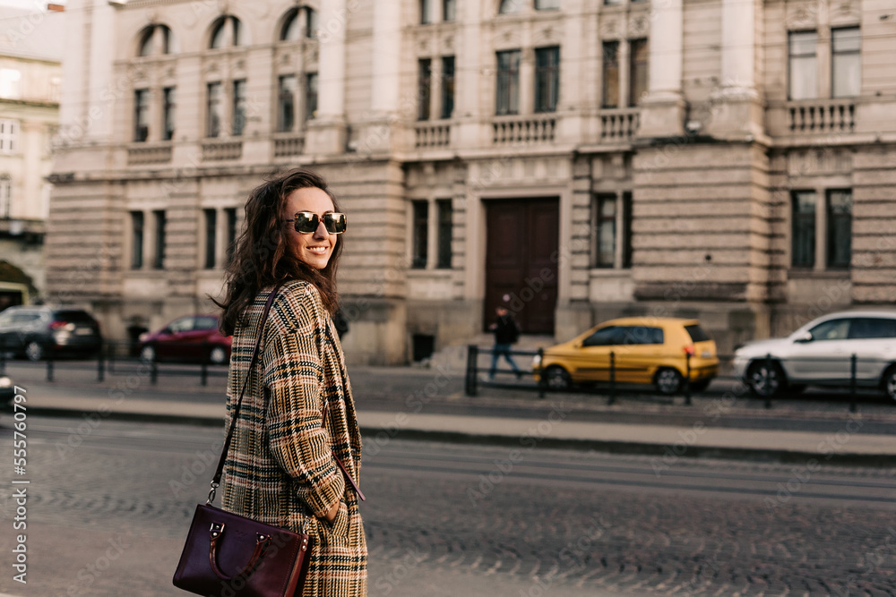 Front view of fashionable woman, with long hair, dressed in stylish coat and having bag on shoulder, looking at the camera while standing against of city building near city street