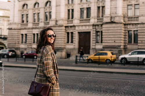 Front view of fashionable woman, with long hair, dressed in stylish coat and having bag on shoulder, looking at the camera while standing against of city building near city street