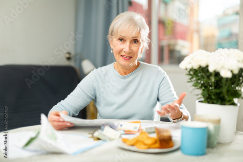 Senior woman sitting at table and looking in camera while counting her budget with bills and documents.