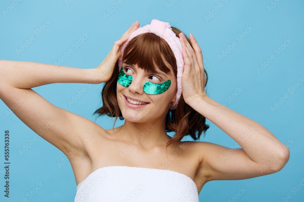 horizontal close beauty photo of a pleasant, joyful woman with clear skin standing on a blue background with colored, shiny patches under her eyes, holding her soft rim with her hands horizontal close beauty photo of a pleasant, joyful woman with clear skin standing on a blue background with colored, shiny patches under her eyes, holding her soft rim with her hands