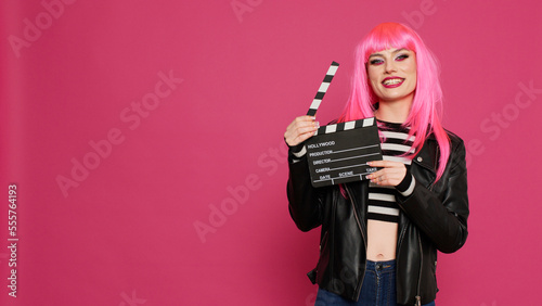 Cheerful female videomaker holding film slate and saying action, preparing to record video montage or footage. Young beautiful person with filming equipment, filmmaker over background.