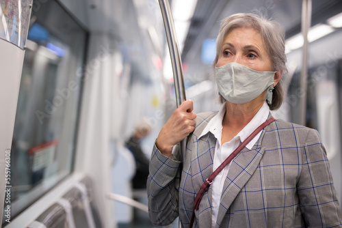 Mature European woman in face mask standing in subway train and holding handrail.