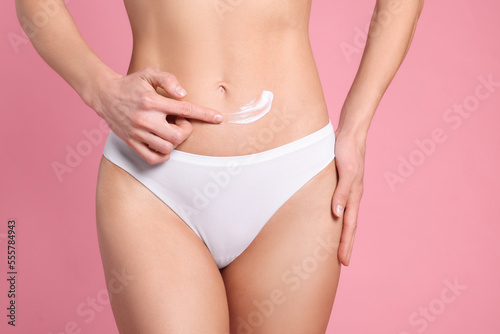 Woman applying body cream onto her belly against pink background, closeup