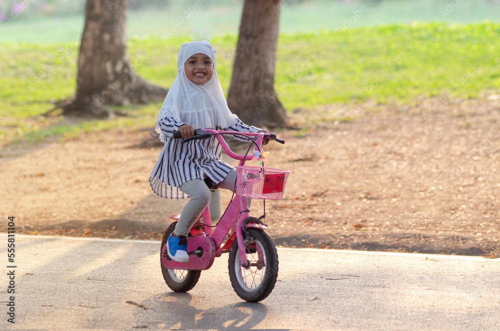 Smiling Muslim little girl riding bicycle in the park, in autumn park at sunset, looking at camera Smiling Muslim little girl riding bicycle in the park, in autumn park at sunset, looking at camera