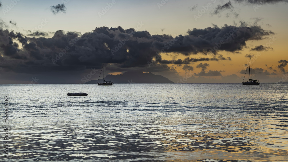 Evening twilight in the tropics. Boats are visible in the calm ocean. The sky is highlighted in orange. The island on the horizon is hidden in picturesque clouds. Seychelles. Mahe
