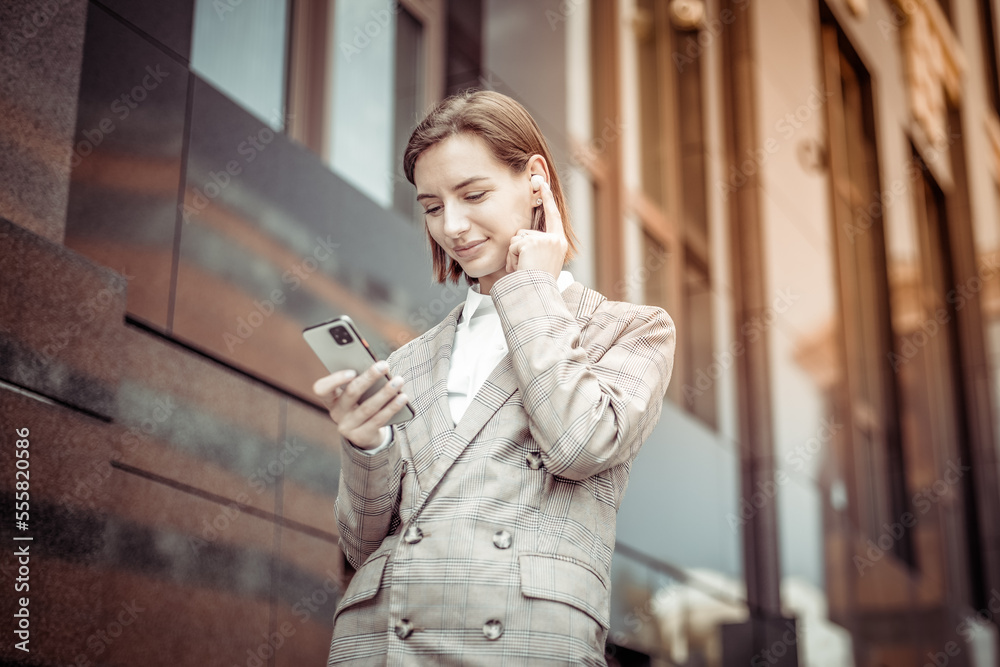 Fototapeta premium Young modern woman in a jacket touching the earphone while looking into the smartphone screen