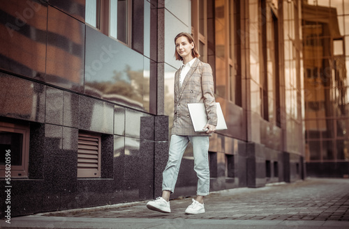 Informal, young business woman with a laptop walks through the city on the background of a business building
