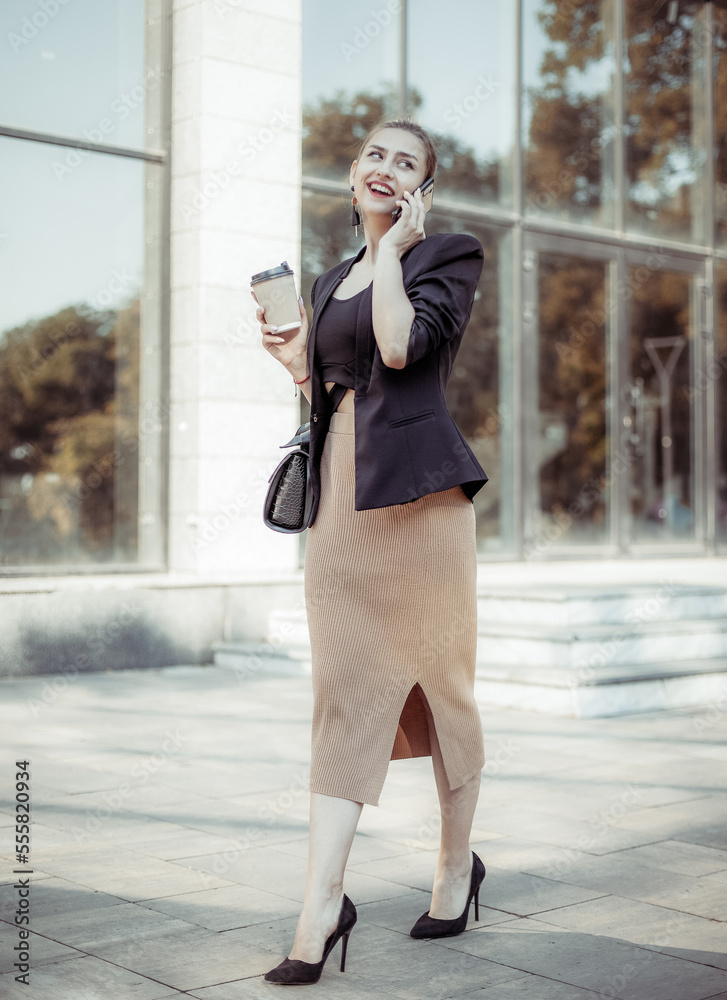 Young business woman talking on phone and drinking coffee on the go outdoors